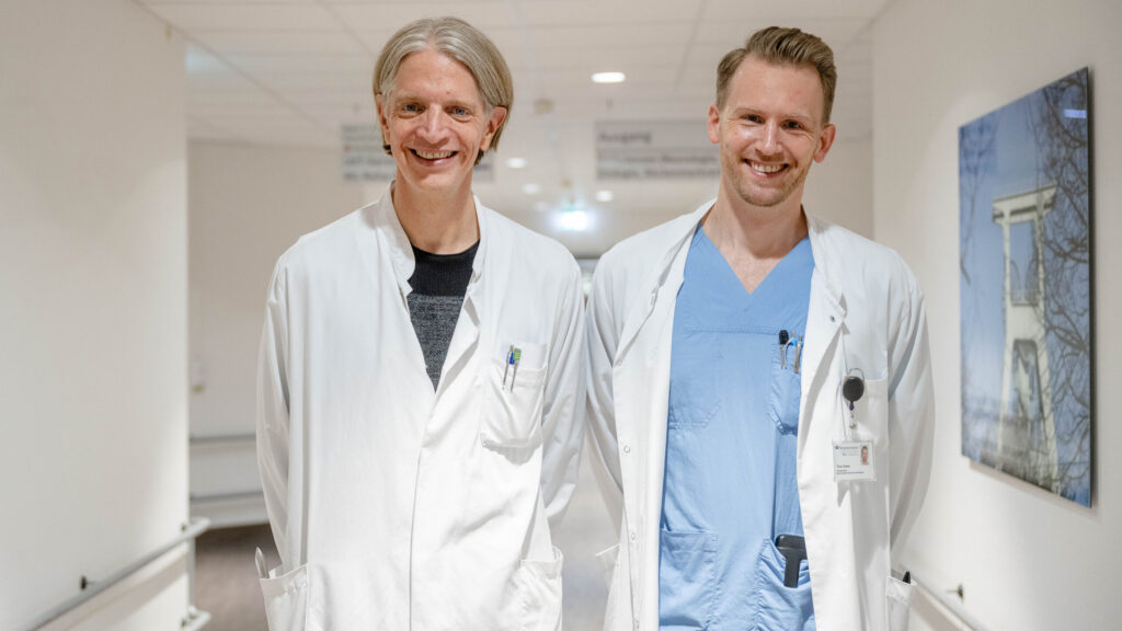 Foto von Burkhard Pleger und Timo Dreier in einem Krankenhausflur. Beide tragen Arztkittel. Ein Bild an der Wand zeigt einen Förderturm.